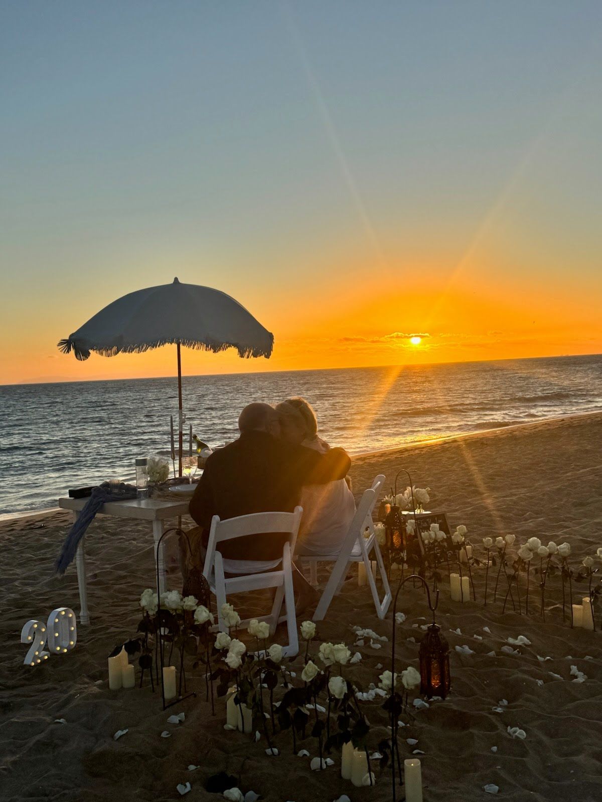 A couple, seen from behind, sits at a small table on the beach at sunset, surrounded by white roses, candles and lanterns under a fringed umbrella.