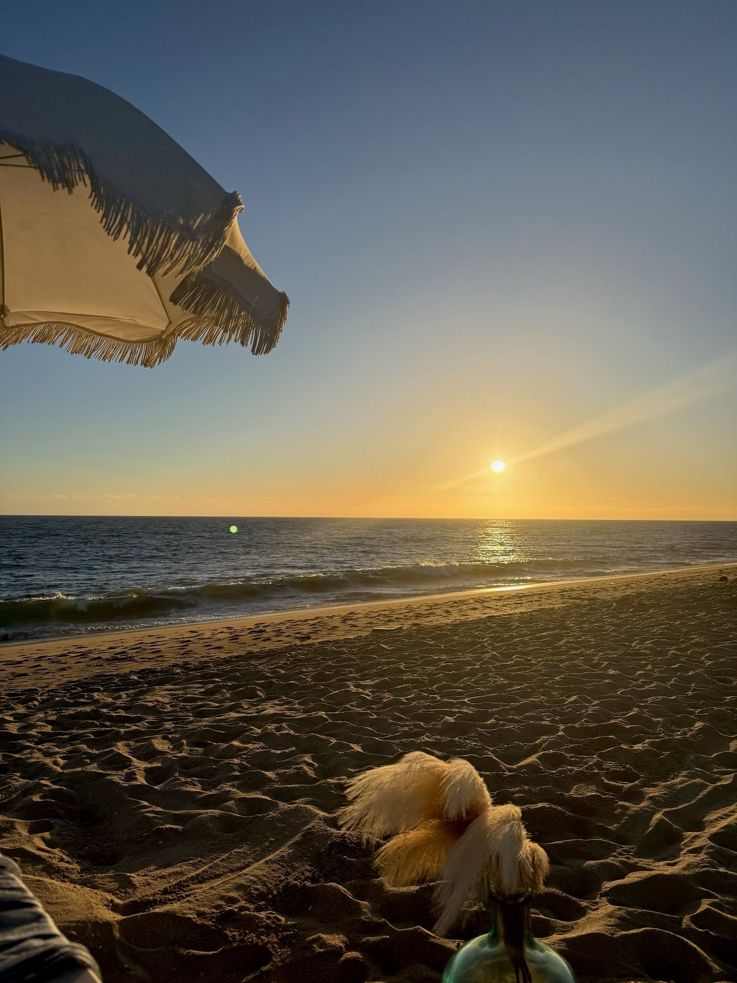 The sun setting over the ocean, framed by the edge of a fringed beach umbrella, with soft pampas grass in the foreground.